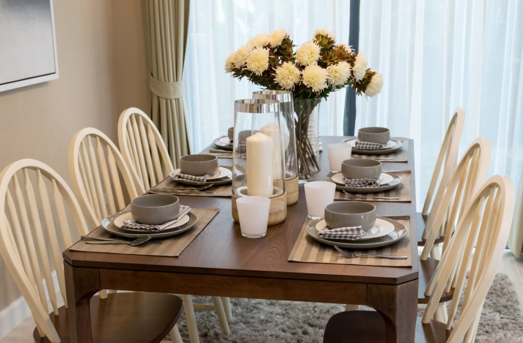 A wooden dining table with 3 chairs on opposite sides, plates on the table, flowers in a vase and 2 candle stands.