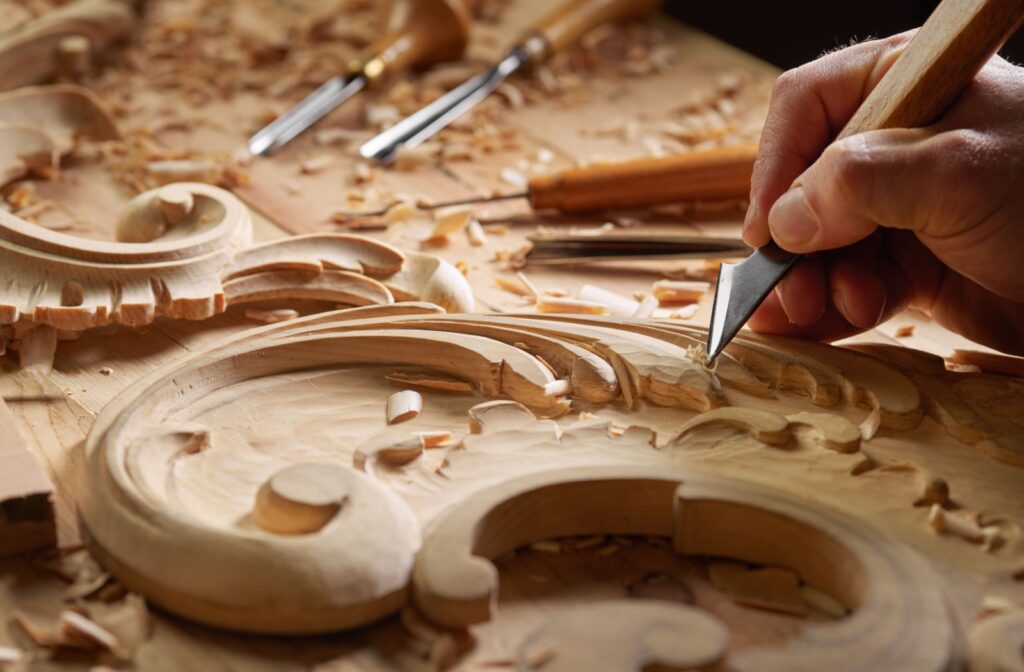 A close-up of a furniture maker carving ornate wing and feather designs into a piece of wood.