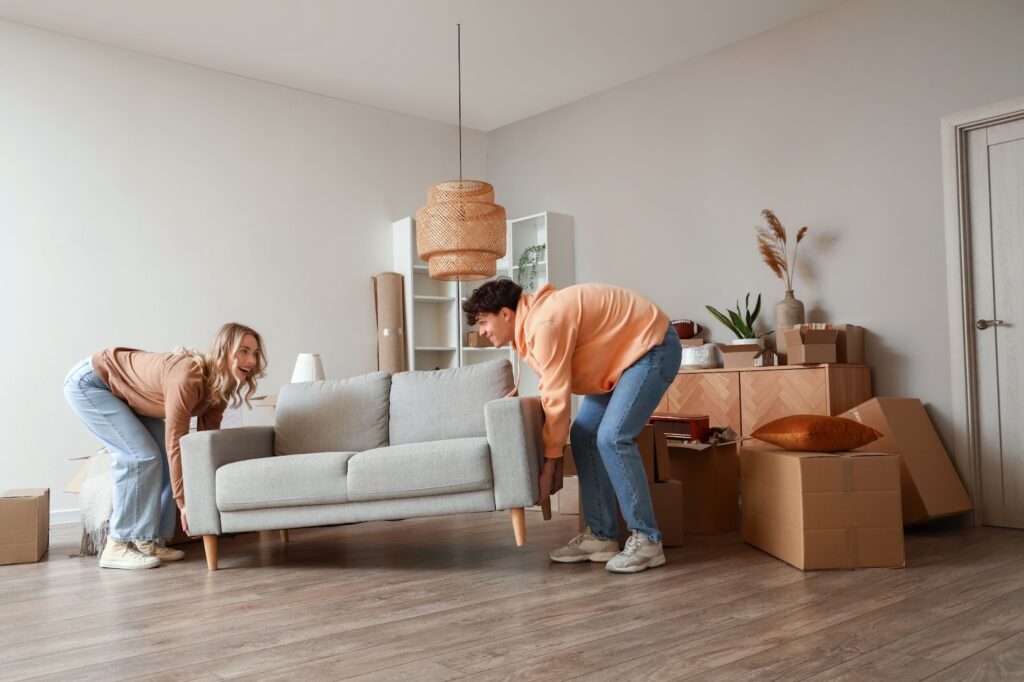 2 people moving a small loveseat in a living room filled with boxes and other new furniture pieces.